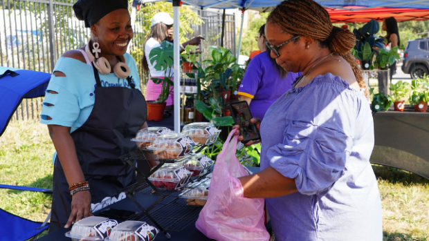 One woman buys produce from another woman under a farmer's market tent.