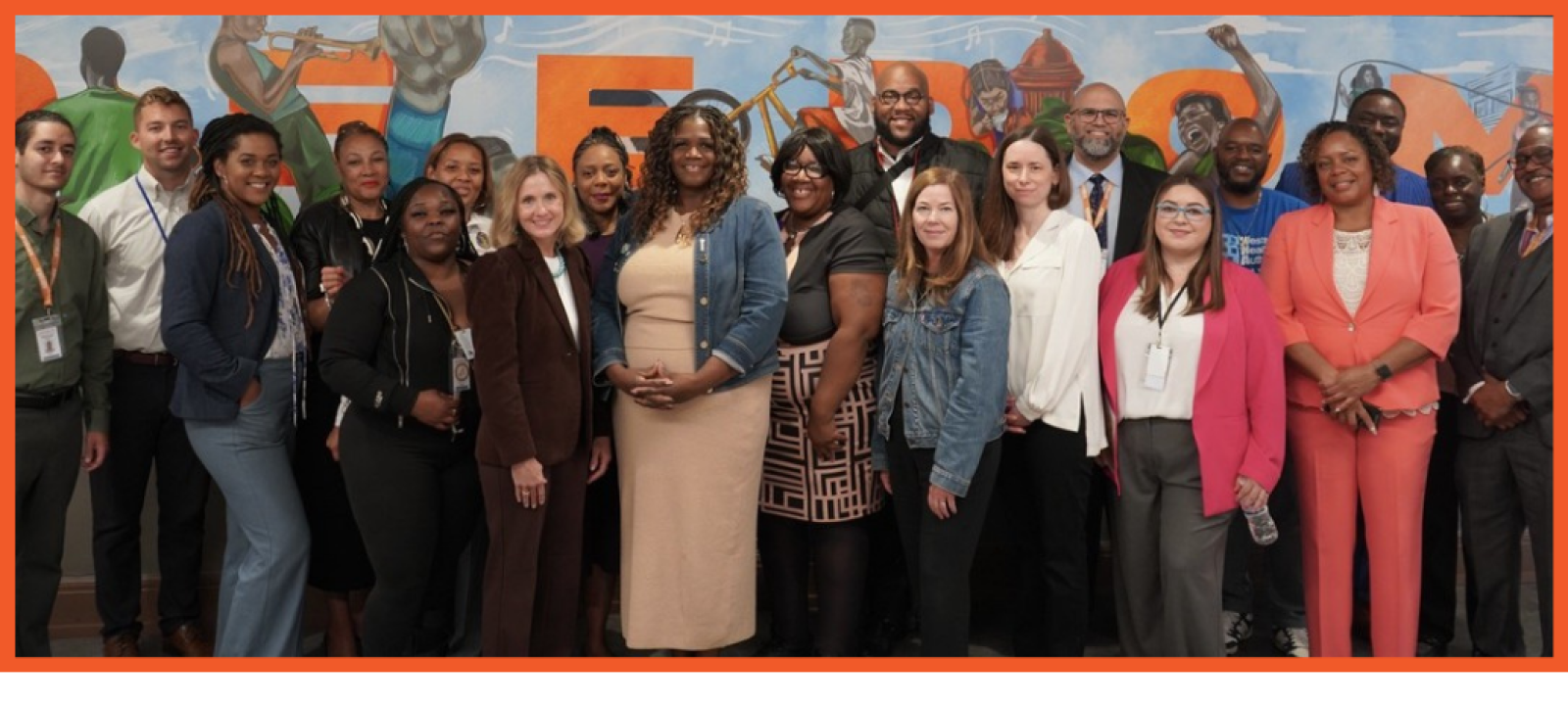 A large group of colleagues and community members stand in front of a colorful mural that reads "freedom"