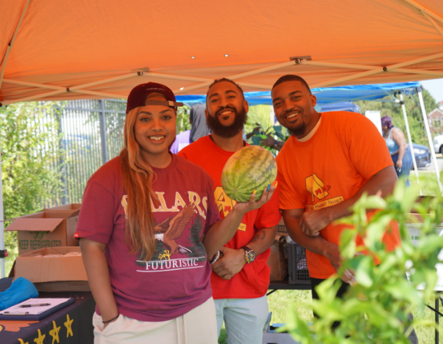 Three young adults smile for the camera while selling produce at a farmer's market.