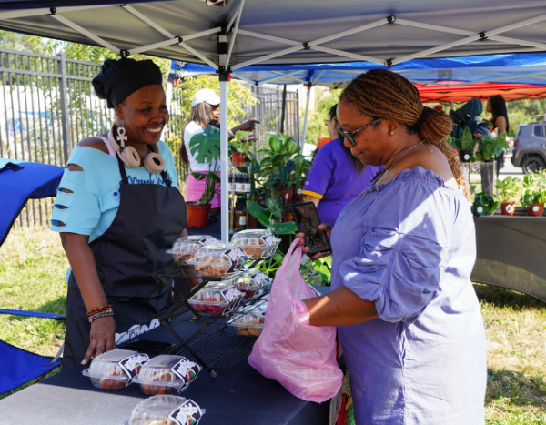 One woman buys produce from another woman under a farmer's market tent.